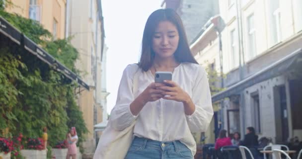 Étudiante joyeuse rentrant à la maison après une dure journée d'étude et en utilisant un smartphone. Jeune femme asiatique marchant dans la rue de la ville, message texte et sourire. Communication, concept technologique. — Video