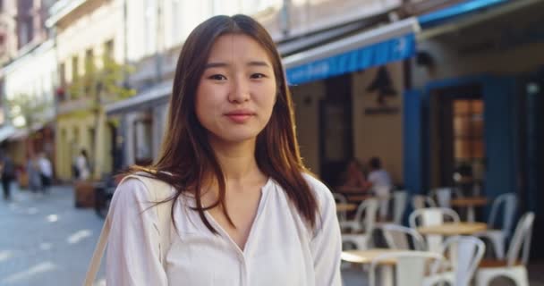 Portrét krásné stylové asijské ženy dívající se a usmívající se na kameru ve staré evropské ulici. Sebevědomá mladá dáma v neformální bílé košili. Městské zázemí. Řada Real People. — Stock video