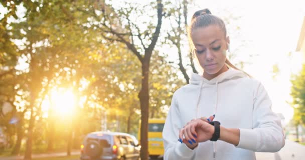 Attraente donna caucasica in auricolare ascoltare musica e impostare stop watch. Adulto atleta femminile tempismo jogging e cercando di battere il record in strada. Sport, allenamento, concetto di allenamento. — Video Stock