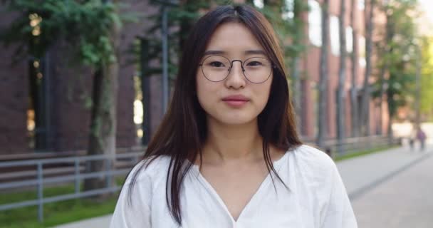 Porträtt av chaming asiatisk kvinna i glasögon tittar på kameran amd leende i urban affärsdistrikt. Snygg ung mörkhårig dam står utanför modern byggnad på stadens gator. Människobegreppet. — Stockvideo