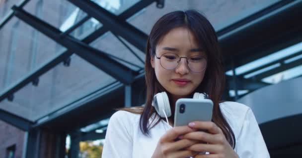 Portrait of good-looking Asian lady in glasses standing outside modern office building. Young woman with headphones using digital smartphone, scrolling news feed, messaging and browsing on Internet. — Stock Video