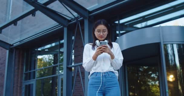 Jovem mulher de cabelos escuros com fones de ouvido em pé no distrito de edifícios de negócios. Muito asiático feminino mensagens de texto, mensagens e navegação no smartphone na rua da cidade. Conceito de tecnologia. — Vídeo de Stock