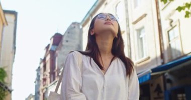 Çevre çantalı çekici genç bir kadın modern şehir meydanında yürüyor. Bardaklı Asyalı kadın turist binalara bakıyor, düşünüyor ve sokakta içiyor. Seyahat, yürüyüş konsepti.