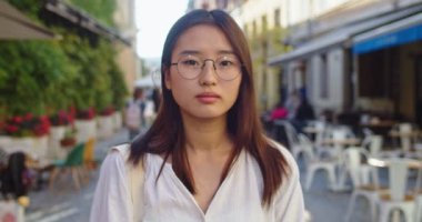Gözlüklü güzel Asyalı kadın portresi kameraya bakıyor ve dışarıda gülümsüyor. Bahar günü şehir caddesinde duran beyaz tişörtlü genç bir bayan. Boş zaman, yürüyüş konsepti.