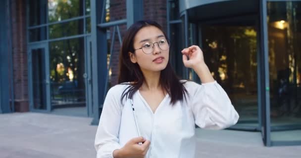 Pěkně vypadající pracovnice opouštějící moderní kancelářskou budovu v obchodní čtvrti. Mladá Asiatka v brýlích držící notebook a procházející se v městské ulici. Práce, podnikání. — Stock video