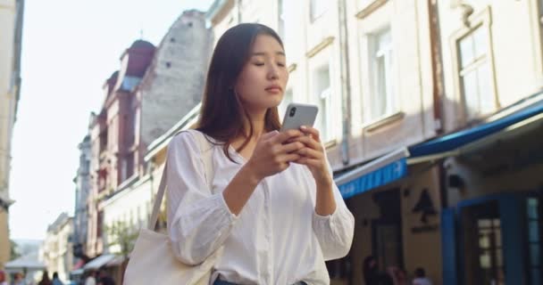 混乱した女性観光客は彼女の方法を失い、スマートフォンアプリでアドレスを検索しようとしています。目的地を見つけるために地図を探している若いアジアの女性。旅、旅、テクノロジーの概念. — ストック動画