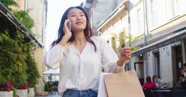 Mujer rizada sonriente con gafas de sol de moda camina por la calle central de la ciudad y utiliza su teléfono. Mujer bonita de verano en chaqueta blanca camina por la calle mirando su teléfono móvil — Vídeo de stock