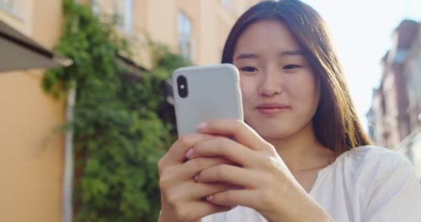 Porträt einer lächelnden jungen Frau, die ihr Smartphone in der Hand hält und eine lustige Nachricht liest. Asiatische Mädchen chatten mit Freunden, scrollen Feeds, stöbern und lachen. Soziale Medien, Kommunikationskonzept. — Stockvideo