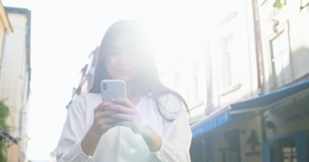 Charmante étudiante utilisant un smartphone et debout dans la rue urbaine. Jeune femme asiatique textos, navigation et sourire. Médias sociaux, technologie, concept de communication. — Video