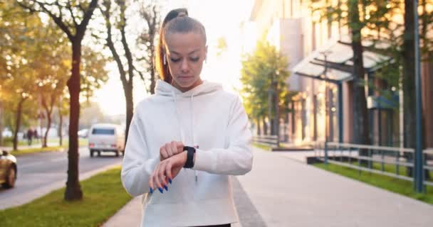 Sterke vrouwelijke atleet met behulp van stop watch to time joggen op slimme digitale gadget. Jonge blanke vrouw die een apparaat installeert en loopt langs moderne gebouwen in de straat. Sport, gezondheidsconcept. — Stockvideo