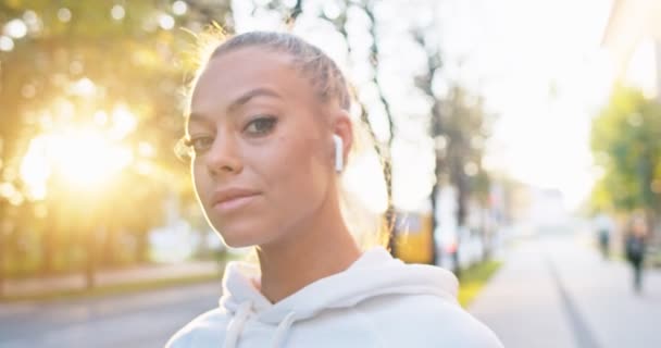 Retrato de atleta feminina confiante com fones de ouvido ouvindo música na rua. Mulher caucasiana jovem em sportswear em pé na área do parque e olhando para a câmera. Pessoas, treino, conceito de jogging. — Vídeo de Stock