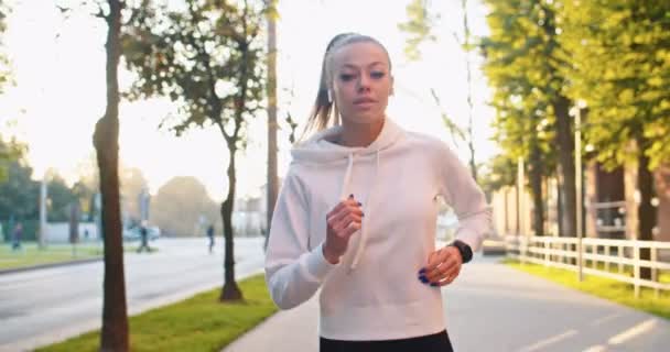 Belle femme caucasienne courant et regardant à la caméra dans la rue de la ville. Athlète féminine adulte en tenue de sport, s'entraînant à l'extérieur le matin. Santé, sport, mode de vie actif concept. — Video