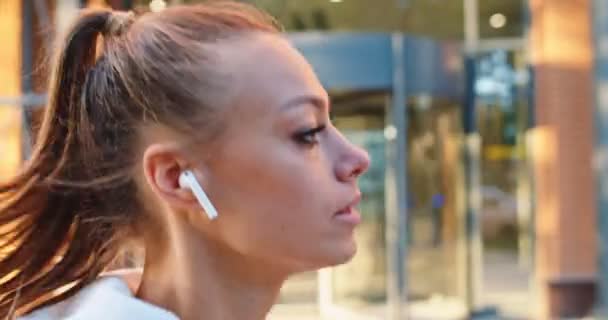 Side view of good-looking Caucasian woman training, jogging, working out in city urban street. Young female athlete in earphones running along modern business buildings. Sport, health concept. — Stock Video