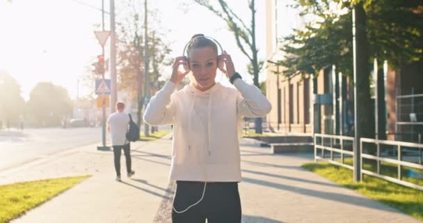 Mujer caucásica fuerte haciendo ejercicio, entrenando en el día cálido de verano. Guapa atleta femenina que se pone auriculares para escuchar música y correr, trotando en la calle de la ciudad. Deportes, salud, concepto de fitness. — Vídeos de Stock