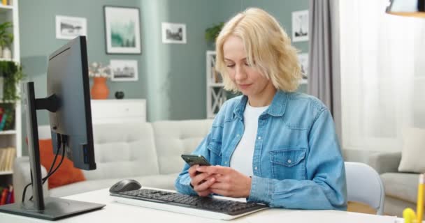Кавказька вродлива молода жінка сидить за столом у сучасній кімнаті і друкує текстове повідомлення на смартфоні і дивиться онлайн на комп'ютер, який працює вдома. Портрет красивої жінки, що друкує на пристрої — стокове відео