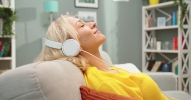 Beyaz kulaklıklarla şarkıların tadını çıkaran, evinde, kanepede dinlenen, mutlu, rahat, beyaz, genç, güzel bir kadın. Oturma odasında müzik dinleyen güzel bir kadının yan görüntüsü