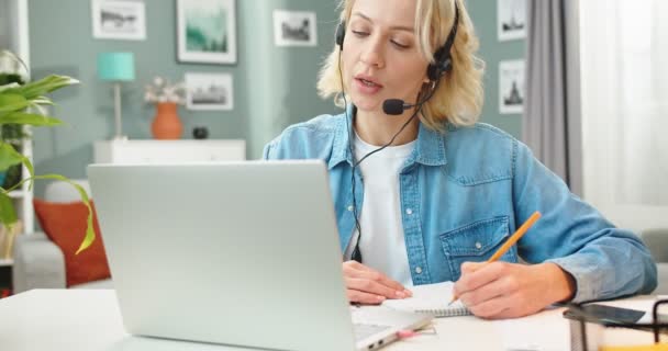 Primer plano de Caucásico joven hermosa rubia estudiante videochat en el ordenador portátil hablando en la lección en línea y tomando notas sentado en el escritorio en la sala de estar, la educación en Internet, webinar — Vídeo de stock