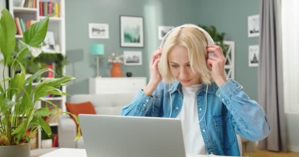 Retrato de la alegre joven hermosa mujer caucásica sentada en el escritorio en la habitación en casa escuchando música canción en auriculares blancos usando el ordenador portátil que se divierte bailando de buen humor, concepto de ocio — Vídeos de Stock