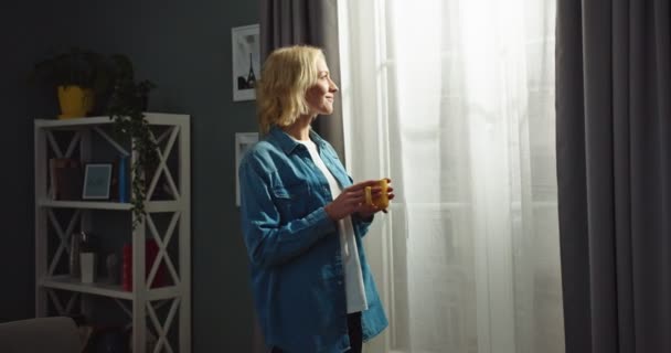 Portret van vrolijke mooie blanke jonge blonde vrouw die in de woonkamer in het appartement staat, warme koffie of thee drinkt en in het raam kijkt met een dromerig gezicht. Vrije tijd, huiselijk thuis — Stockvideo