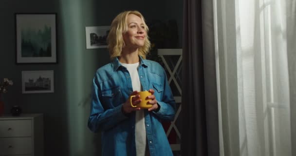 Retrato de joven alegre hermosa mujer rubia caucásica de pie en la sala de estar oscura en la casa, mirando en la ventana con la cara feliz de ensueño mientras sostiene la taza de bebida caliente, concepto de hogar — Vídeos de Stock