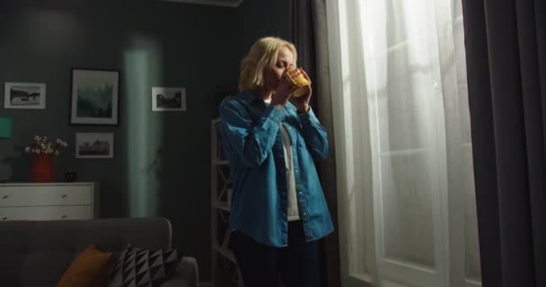 Caucasian young beautiful blonde young woman standing in dark cozy living room in apartment, looking in window with dreamy happy face while sipping hot drink, coffee lover, rest concept — Stock Video
