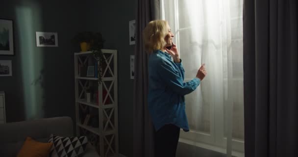 Kaukasisk ung vacker glad ung kvinna som står i mörkt vardagsrum i lägenheten, tittar i fönstret medan du talar på smartphone med glada ansikte. En söt kvinna ringer på mobilen hemma. — Stockvideo