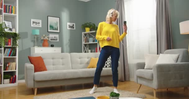 Kaukázusi boldog pozitív gyönyörű nő táncol és ugrál a modern nappaliban, miközben zenét hallgat fülhallgatóban okostelefonról. Nagyon vidám nő, jól érzi magát otthon. — Stock videók