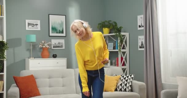 Porträtt av vit glad vacker blond kvinna dansar ha kul i moderna vardagsrum i lägenhet medan du lyssnar på fav sång i hörlurar från smartphone. Fritid, avslappning koncept — Stockvideo