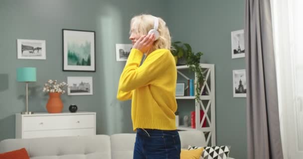 Retrato de la alegre joven caucásica hermosa mujer bailando en la sala de estar moderna en el apartamento mientras escucha música en los auriculares en estado de ánimo positivo. alegre rubia hembra tener divertido disfrutando de canción — Vídeos de Stock