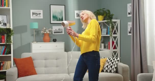 Mladá běloška krásná šťastná atraktivní žena ve žlutém svetru tančí v moderním obývacím pokoji v bytě, zatímco zpívá do fénu v pozitivní náladě. Veselá blondýnka se baví doma