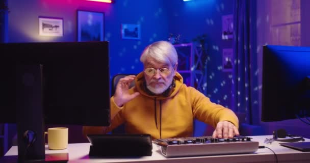 Portret szczęśliwego starszego DJ-a grającego na sprzęcie w domu nagrywającego nowe studio muzyczne. Stary samiec uczy się pisać kreatywną atmosferę muzyki w pokoju na komputerze w kolorowe neony — Wideo stockowe