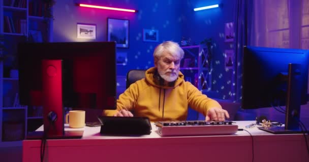 Портрет счастливого старшего человека ди-джея в наушниках работает на микшерной консоли, создавая музыку на оборудовании, сидящем за столом дома, печатая на компьютере в неоновом свете, старый профессиональный ди-джей создает звуковой бит — стоковое видео