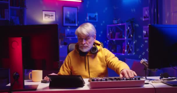 Portré kaukázusi öreg férfi profi DJ dolgozik a házban neon fények, zeneszerző streaming online számítógépen. Művész dolgozik otthon rekord stúdió segítségével felületvezérlő pult kiegyenlítő mixer — Stock videók