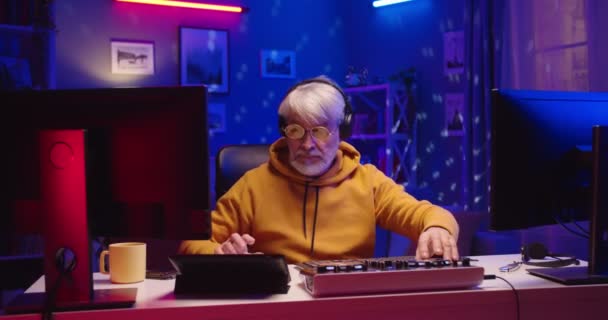 Happy funny stylish old male DJ working at home studio making music on mixer console streaming online on computer listening to sound in headphones and spinning on chair at home in neon lights portrait — Stock Video