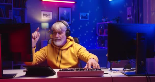 Portrait of happy Caucasian senior grey-haired male DJ in headphones using mixing console and computer while creating dance music in home recording studio with colorful neon light, job concept — Stock Video