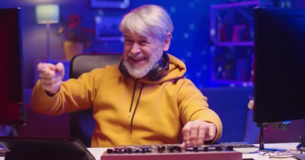 Közelkép portré pozitív örömteli kaukázusi öregember DJ ül íróasztal a szobában neon fényben dolgozik professzionális mixer konzol és számítógép létrehozása zene élvezi folyamat, hobbi koncepció — Stock videók