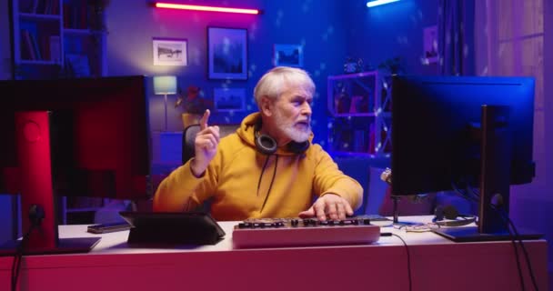 Gioioso pensionato caucasico senior man DJ seduto alla scrivania in camera a riprodurre musica a casa studio in luce al neon, DJ professionista che lavora su mixer console musica in streaming utilizzando il computer guardando i monitor — Video Stock