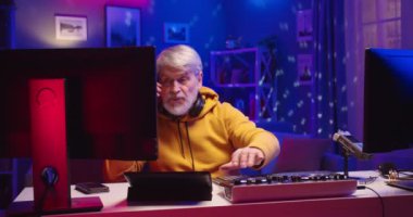 Kafkas tarzı, yaşlı, gri saçlı DJ ev stüdyosunun neon ışığı altında müzik çalıyor karantina sırasında profesyonel mikser konsolu üzerinde çalışıyor ve akıllı telefondan masada oturuyor.