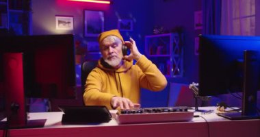 Beyaz saçlı, sarı şapkalı, şık erkek DJ. Ev kayıt stüdyosundaki elektronik müzik setinde profesyonel mikser konsolu, portre, vinil plaka, hobi konsepti üzerinde çalışıyor.