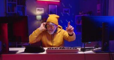 Beyaz tenli emekli erkek DJ 'in portresi. Sarı süveterli, elektronik müzik kaydediyor. Neon ışıkta, profesyonel mikser konsolunu kullanıyor.