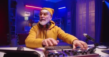 Beyaz sakallı, mutlu, sarı kazaklı DJ 'in portresi konsol karıştırıcısı üzerinde çalışıyor. Plastik plaka ekipmanlarıyla yeni müziği karıştırıyor. Ev stüdyolarının neon ışıklarındaki masasında oturuyor.