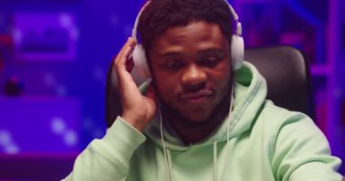 Afro-Amerikan yakışıklı genç ve neşeli DJ 'in yakın portresi. Ev kayıt stüdyosunda oturmuş kulaklıkla ses dinliyor. Kulüp elektronik müziğini neon ışıkta, hobide, iş konseptinde karıştırıyor.