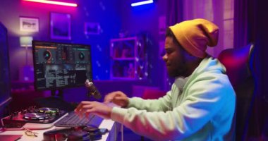 Şık, genç, Afro-Amerikan DJ 'i akıllı telefon kullanarak ses yaratıyor mikser konsolunda müzik karıştırıyor kulaklıklarda müzik dinliyor neon ışıklarda, ses mühendisi, kenar görüşlü.