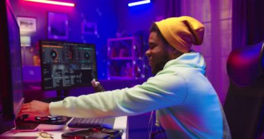 Neşeli Afrikalı Amerikalı genç ses mühendisi müzik besteleyerek mikser konsolunda ses yaratıyor kulaklıklarda müzik dinliyor karanlık odada neon ışıkları, yan görüş, DJ konsepti.