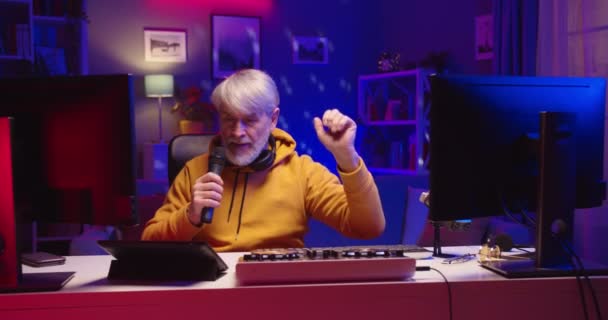 Portré idősebb fehér öreg férfi DJ ül a szobában neon fény otthon streaming élő zene karanténban beszél mikrofonban, MC beszél, professzionális DJ konzol mixer berendezések — Stock videók