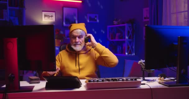 Stylish senior Caucasian bearded retired man in yellow hat streaming live DJ set at home working on professional mixer console equipment, music streaming podcast, portrait, work, hobby concept — Stock Video