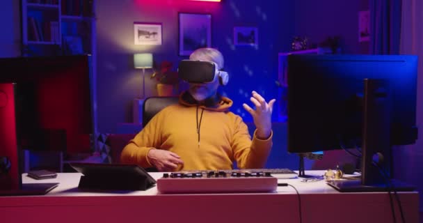 Portrait of old Caucasian pensioner male DJ sitting at table in dark room with neon light in home recording studio wearing VR glasses playing guitar in virtual reality, modern technology music concept — Stock Video