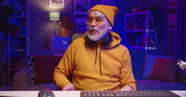 Portrait of modern stylish Caucasian bearded old man DJ sitting in dark room in neon lights with headphones mixing new music, senior male dj in positive mood dancing on chair, electronic music — Stock Video