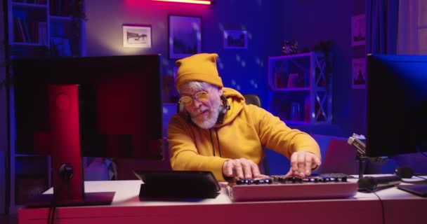 Stylish Caucasian senior bearded male DJ in yellow sweater streaming or recording electronic music at home studio with neon light working on professional mixer console mixing on vinyl plate, portrait — Stock Video