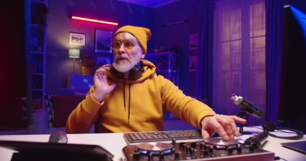 Großaufnahme von kaukasischen stilvollen Rentner männlichen DJ sitzt am Tisch in dunklen Raum mit Neonlicht zu Hause Studio arbeiten an Mischpult neue Musik auf Vinyl-Platte, Sound-Beat-Maker — Stockvideo